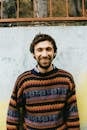 Happy adult bearded male with toothy smile in knitted sweater standing near colorful shabby wall with metal fence above in daylight and looking at camera
