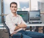 Confident young man seated in a modern office setting with laptops and screens around him.